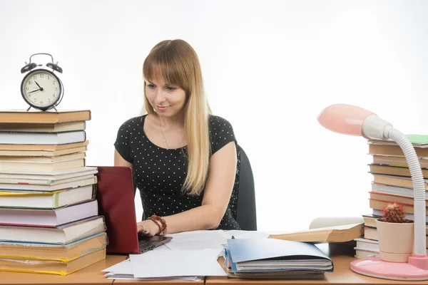 Flickan bakom skrivbordet fylld med böcker leende i en laptop informationsinsamling — Stockfoto