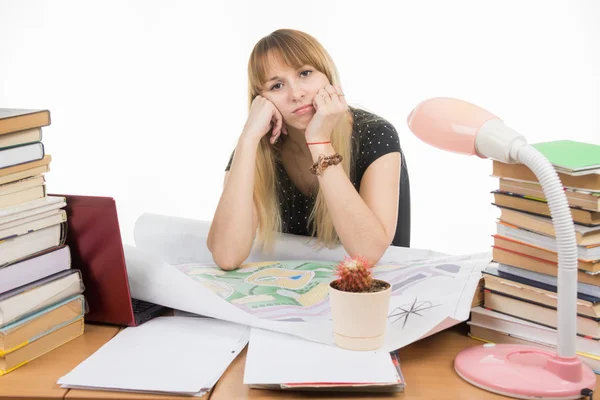 The girl is sad student sitting at a table crammed with books, drawings — Stock Photo, Image