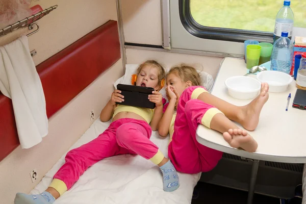 Due bambine si trovano sul treno con i piedi sulla macchina di seconda classe inferiore nello stesso pigiama e guardando tablet — Foto Stock