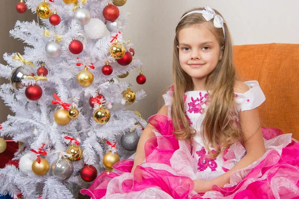 Seven-year girl sitting on a sofa in a beautiful dress the Christmas tree — Stock Photo, Image