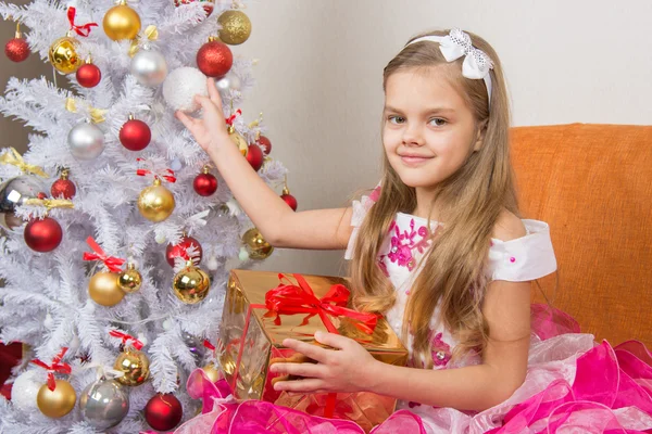 Sjuårig flicka i en vacker klänning sitter med en gåva och håller en jul boll i händer — Stockfoto