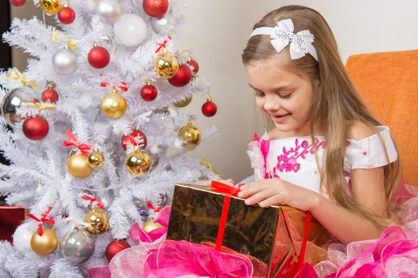 Siete años chica en un hermoso vestido está considerando un regalo sentado en el sofá en el árbol de Navidad —  Fotos de Stock