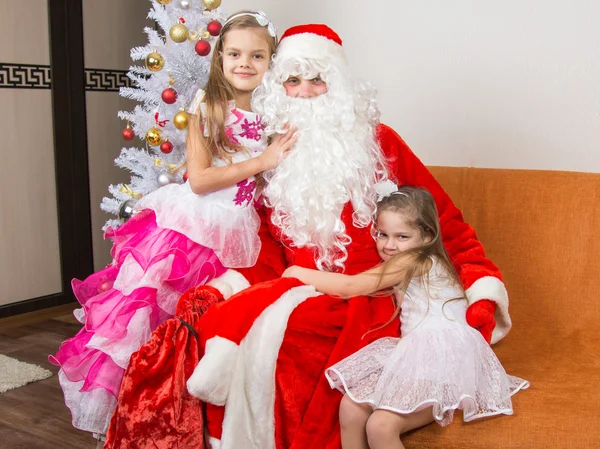 Due ragazze in bei vestiti abbracciano Babbo Natale seduto su un divano — Foto Stock