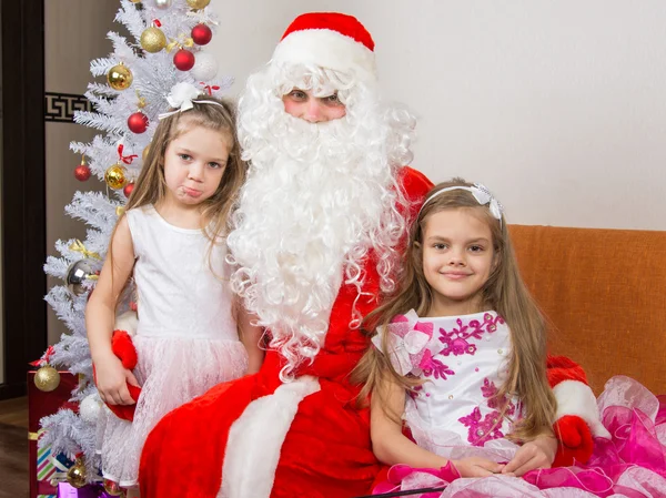 Dwie dziewczyny w piękne sukienki przytulić Santa Claus siedzi na kanapie, jeden z nich trochę niezadowolony — Zdjęcie stockowe