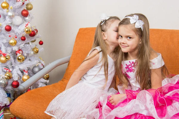 两个女孩穿着漂亮的连衣裙耳语坐在沙发上在圣诞树 图库图片