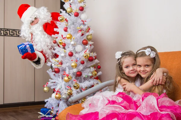 Две девушки обнимаются на диване, Санта Клаус подглядывает из-за деревьев — стоковое фото