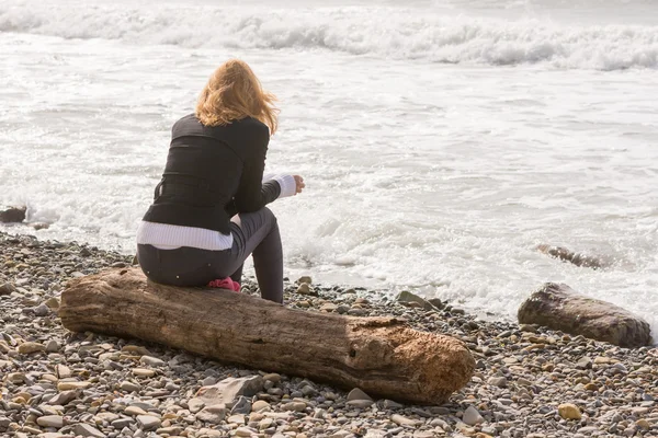 Girl sitting on the log on the coast and looks at sea — Stock Photo, Image