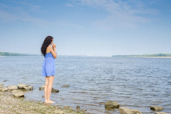 Sad young brunette woman in a blue summer dress standing on the river bank — Stock Photo, Image