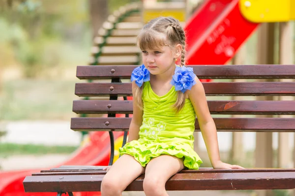 Retrato de niña molesta de cinco años que está sentada en el banco en el fondo del patio de recreo —  Fotos de Stock