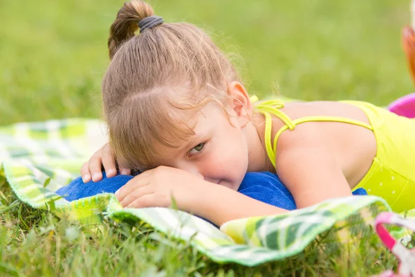 Chica de cinco años se encuentra en una cama en un prado verde y ingeniosamente mira a un lado — Foto de Stock