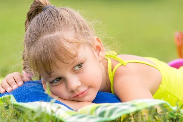Пятилетняя девочка лежит на кровати на зеленом лугу и задумчиво смотрит в сторону — стоковое фото