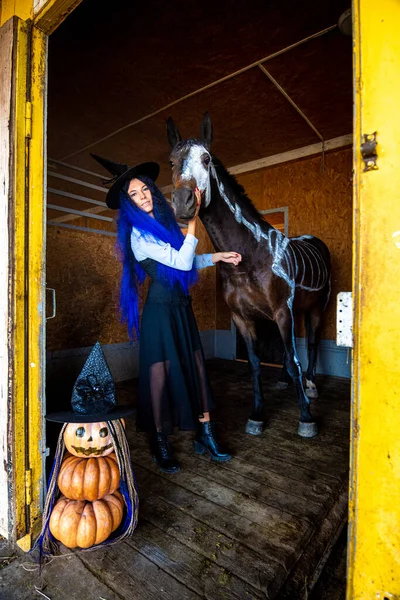 A girl dressed as a witch stands in a corral with a horse on which a skeleton is painted in white paint, in the foreground is an evil figurine of pumpkins