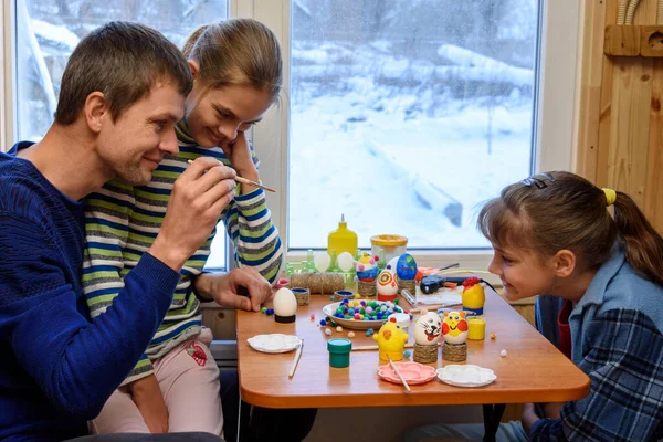 Dad and two daughters paint Easter eggs while sitting at the table by the window