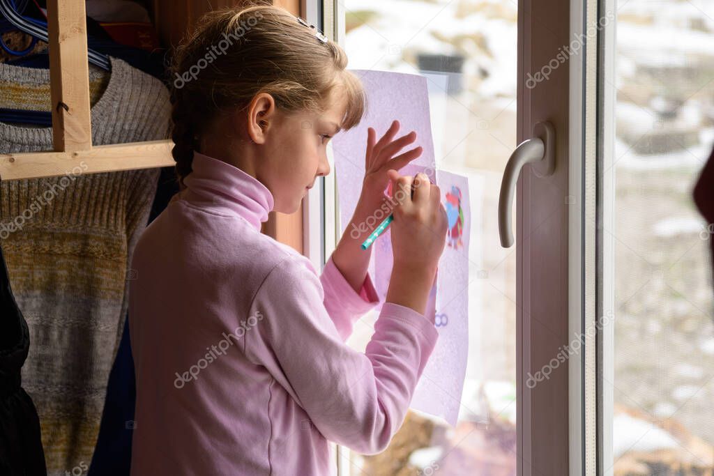The girl draws a picture on a blank sheet by attaching the picture to the window glass