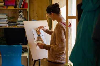 Sanatçı, pencerenin yanındaki küçük bir atölyede sehpada akrilik boyalı bir kedi resmi çiziyor.