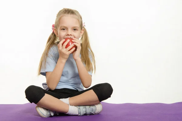 Cheerful girl eating an apple sitting on sport mat — Stock Photo, Image