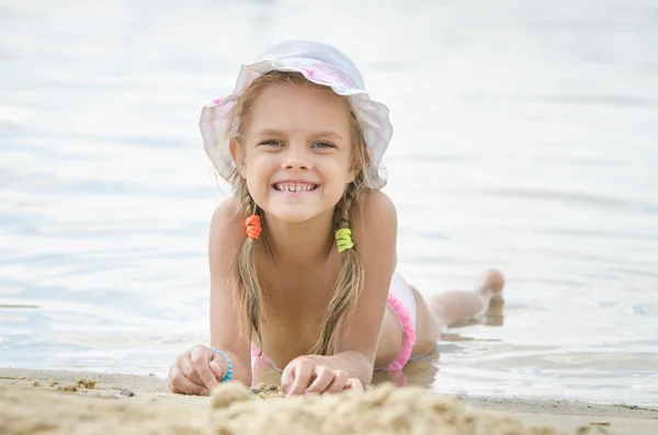 Happy little girl six years lying on the sand on the bank of the river — Stock Photo, Image