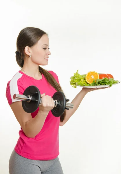 Sportswoman looking at a plate of fruit while holding a dumbbell in the other hand — Stock Fotó