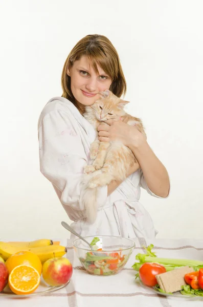 Молодая девушка сидит за столом с вегетарианскими блюдами и держит недовольного кота — стоковое фото
