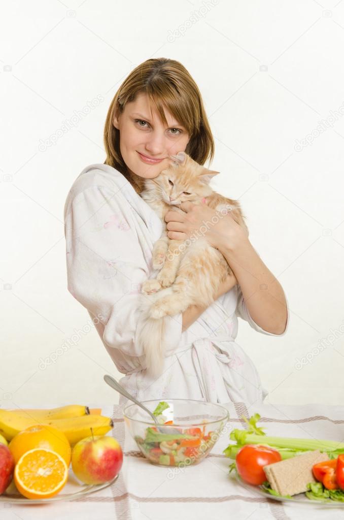 Young girl sitting at the table with vegetarian dishes and holds a disgruntled cat