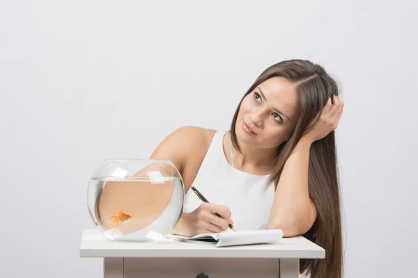 Ragazza premurosamente scrivere in un quaderno, in piedi accanto a un acquario con pesci rossi — Foto Stock