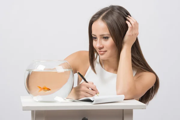 Girl writing in a notebook and looking at a goldfish in an aquarium — Stock Photo, Image