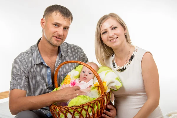 Mamá y papá se sientan en la cama y sostienen a una niña de dos meses en una canasta — Foto de Stock