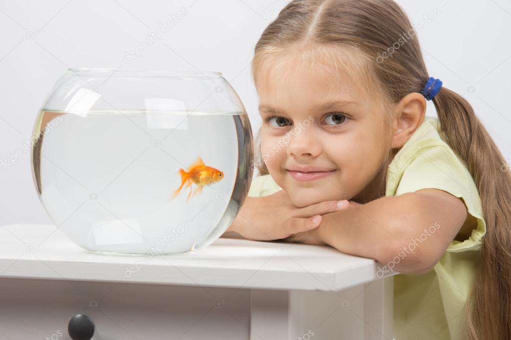 Six year old girl put her head on the handle sitting at the table with an aquarium with goldfish