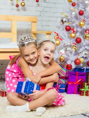 Girl hugging another girl sitting on a rug with a gift at Christmas tree