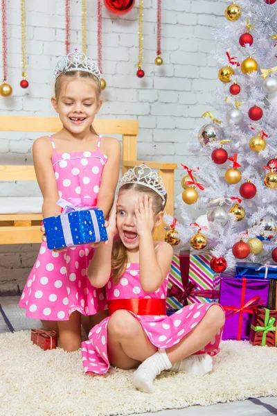 The girl gives a gift to his sister sitting on the rug in front of the Christmas tree — ストック写真