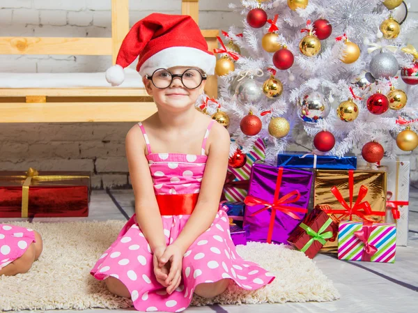 Chica en el sombrero rojo y los divertidos vasos redondos se sienta en la alfombra en los árboles de Navidad —  Fotos de Stock