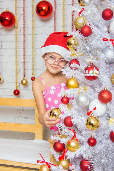 Het meisje in ronde bril en een rode kerst hoed ballen op een besneeuwde nieuwe jaar kerstboom hangt — Stockfoto
