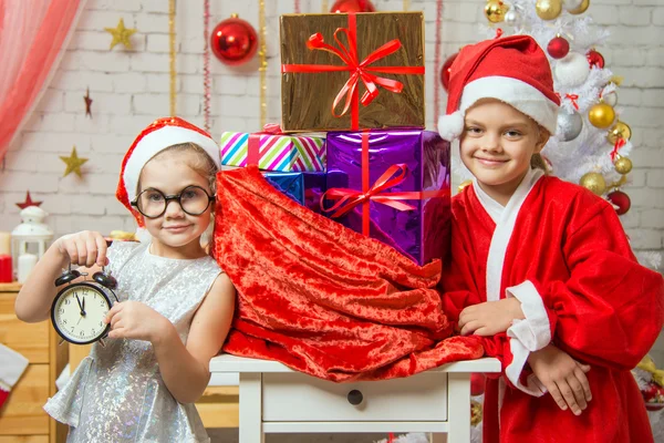 Chica muestra en el reloj, una chica vestida como Santa Claus está de pie en la bolsa con regalos —  Fotos de Stock