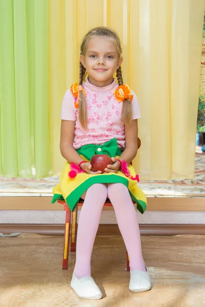 Meisje, zittend op een stoel met een appel in zijn hand op een matinee in de kleuterschool — Stockfoto