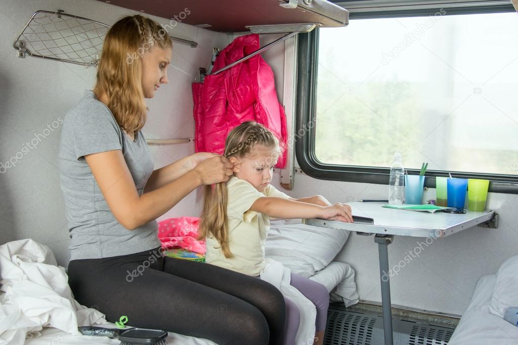 Young mother weave braid of hair daughter on a cot in a train