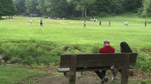 Twee mensen zitten op een bankje in een natuurreservaat kijken jongens spelen bal — Stockvideo