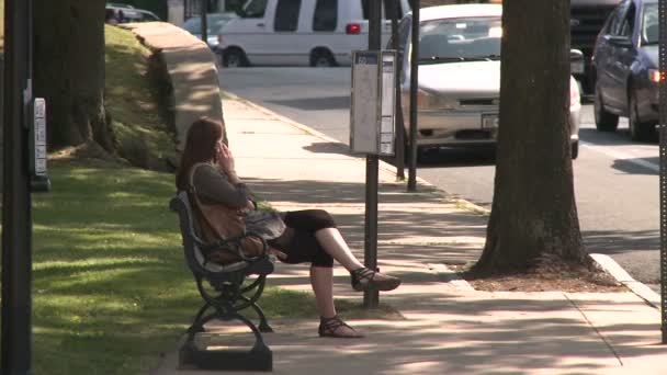 Žena na mobilním telefonu sedí na lavičce podél silnice — Stock video