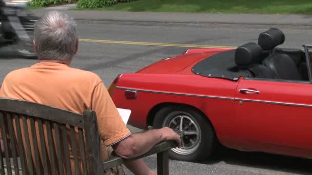 Мужчина, сидящий перед красной спортивной машиной и наблюдающий за проезжающими мотоциклами — стоковое видео