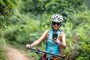 Kadın bisikletçi orman yolunda dağ bisikleti sürerken akıllı telefon kullanır