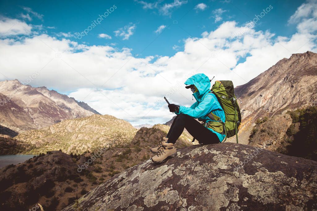Woman backpacker using smartphone sit on beautiful winter mountain top rock