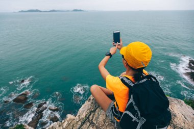 Deniz kenarındaki uçurumun kenarında akıllı telefonla fotoğraf çeken bir kadın yürüyüşçü.