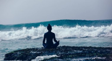 Deniz kenarındaki uçurumun kenarında meditasyon yapan kadın yaklaşan güçlü deniz dalgalarıyla yüzleşiyor.