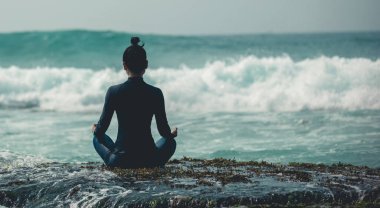 Deniz kenarındaki uçurumun kenarında meditasyon yapan kadın yaklaşan güçlü deniz dalgalarıyla yüzleşiyor.