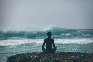 Deniz kenarındaki uçurumun kenarında meditasyon yapan kadın yaklaşan güçlü deniz dalgalarıyla yüzleşiyor.