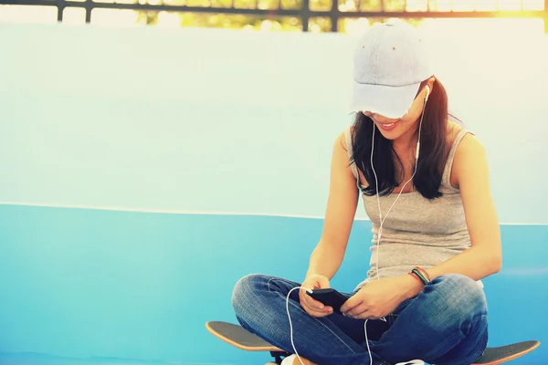 Woman skateboarder sit on skatepark stairs listening music from smart phone mp3 player