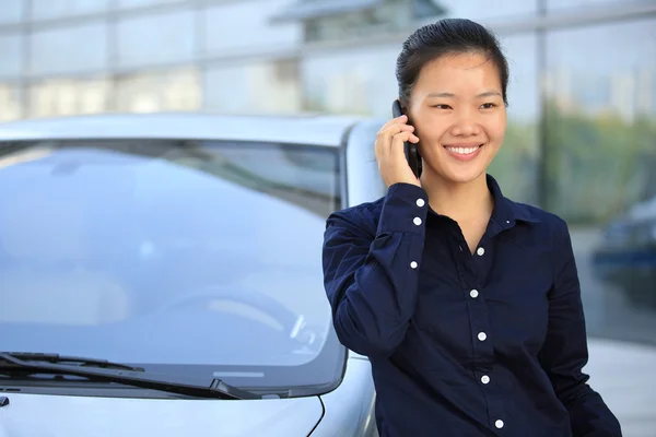 Hermosa mujer de negocios asiática en el teléfono apoyado en el coche fuera del edificio de oficinas — Foto de Stock