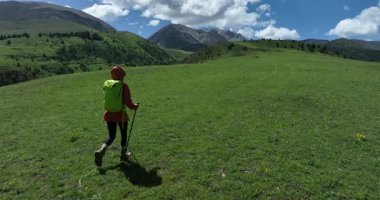 Yürüyüş direkleri olan yürüyüşçü, dağlara doğru güneşli yeşil bir çayırda yürüyor. Yavaş çekim videosu 