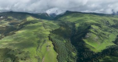 Bereketli, yeşil dağ yamaçları ve bulutlu bir gökyüzünün altındaki ağaç sırtlarıyla çevrili derin bir vadinin havadan görünüşü..