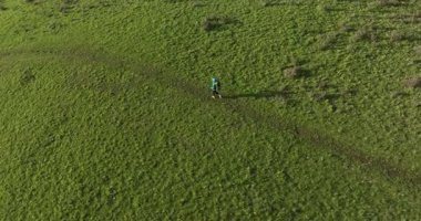 Büyük bir sırt çantalı bir yürüyüşçü arka planda sivri dağ zirveleri olan güneşli, yeşil bir çayırda yürür. Yavaş çekim videosu