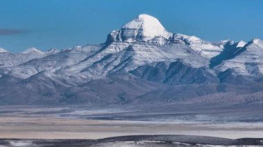 Görkemli, karla kaplı dağ zirvesinin çarpıcı manzarası mavi bir gökyüzünün altında engin ve çorak bir düzlüğün üzerinde yükseliyor..